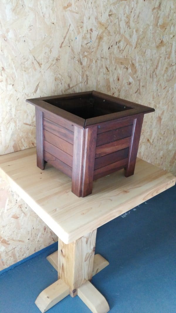 Macetero de madera oscura con forma cuadrada. Su diseño de cuatro patas hace que el interior quede desplazado del suelo.